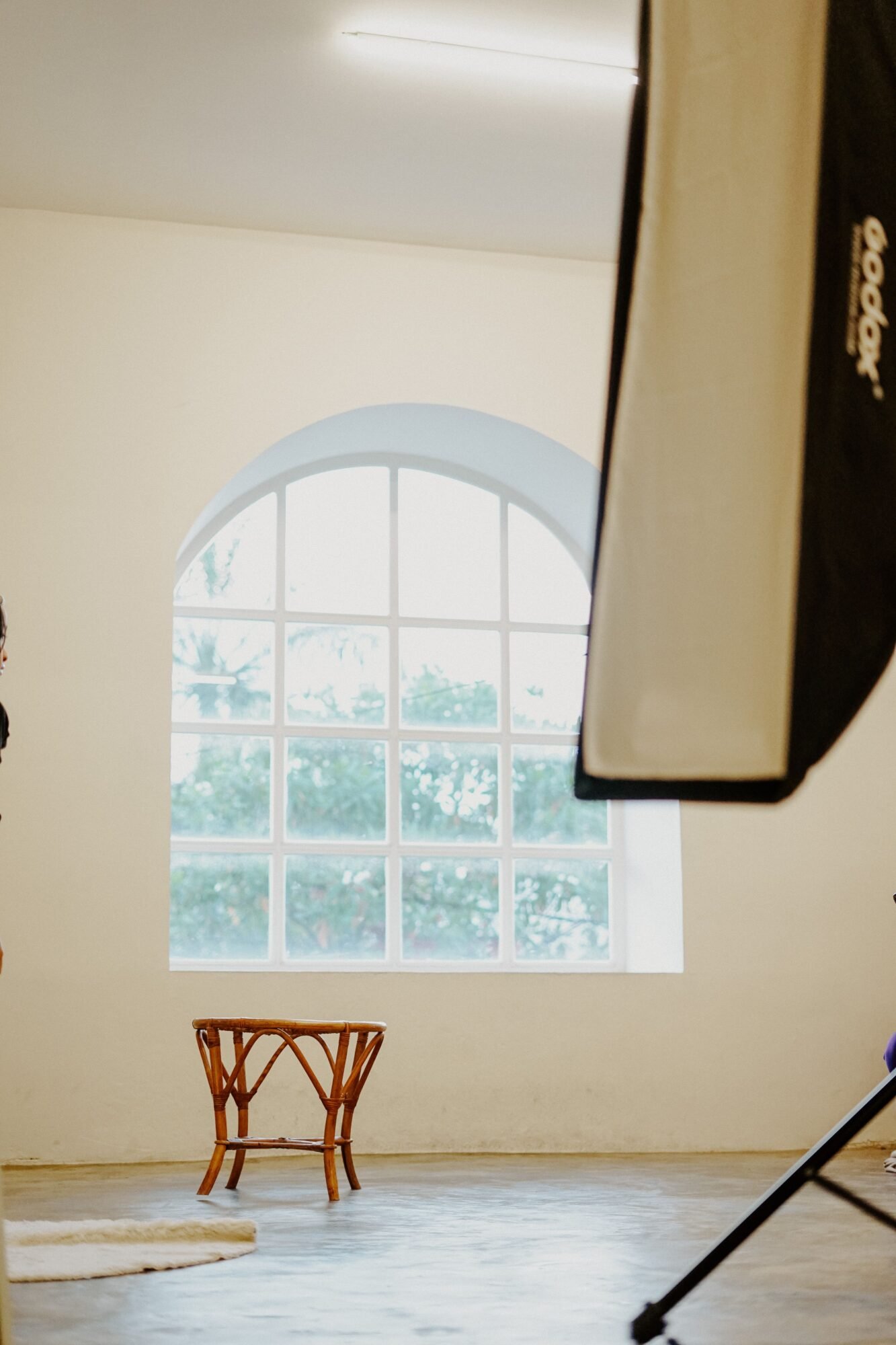 Bright studio with large window, featuring a rattan stool and professional lighting equipment.